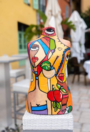 Téléchargez les photos : Menton, France - 9 avril 2024 : Une sculpture moderne d'un torse humain peint avec des motifs d'été - un cocktail, des gens en vêtements d'été, des couleurs chaudes - en image libre de droit