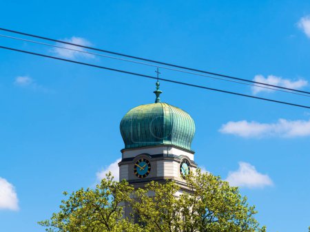 Campanario de la iglesia de cúpula de cobre contra un cielo azul con una pequeña cruz y reloj, enmarcado por exuberantes árboles y líneas eléctricas.