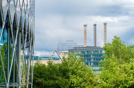 Industrial building with smokestacks, greenery, modern architecture, overcast sky, lattice structure foreground, blending industry with nature.