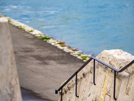 A serene waterside path alongside calm blue Rhine river in Basel, bordered by a stone wall and handrail, creating a peaceful ambiance