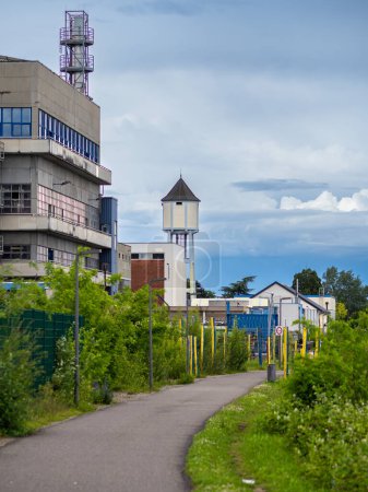 Escena industrial urbana con edificio moderno, torre de agua, vegetación, sendero, postes amarillos y cielo nublado.