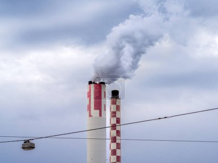 Industrial chimneys in Basel, Switzerland, emitting white smoke, red-and-white striped pattern, utility cable with a street lamp, cloudy sky.