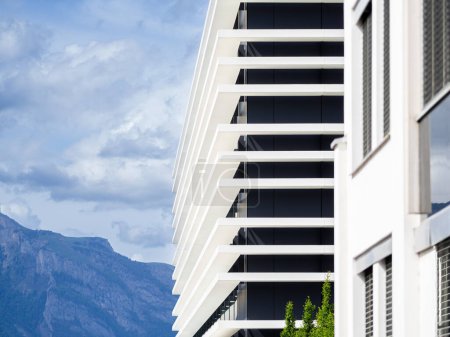 Ein moderner Bau mit weißen, horizontalen Platten und geometrischer Fassade vor felsigen Bergen unter leicht bewölktem Himmel.