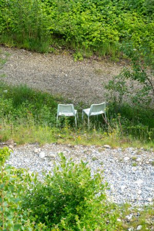Serene setting with two white chairs on grass near rocky path, lush greenery around, perfect for relaxation and conversation.