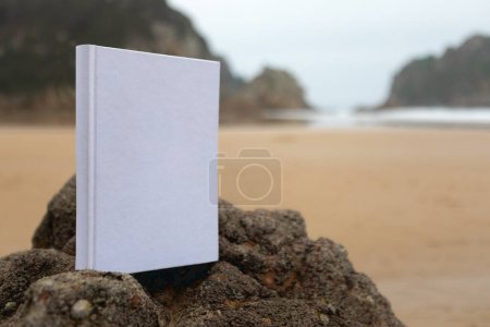 Buch mit weißem Einband ohne Texte oder Zeichnungen im Sand des Strandes an einem bewölkten Tag.
