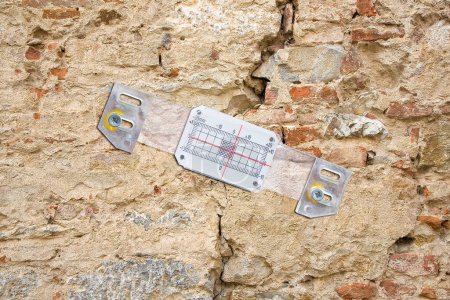 Photo for Deep crack in a damaged old stone wall cause due to subsidence of foundations structural failures with plastic mechanical crack meter designed to measure movement across surface cracks and joints - Royalty Free Image