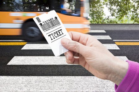 Mano tiene un billete de autobús - concepto de pago de autobús contra un cruce peatonal con el autobús que viene