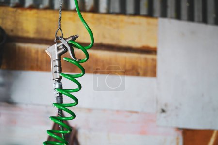 Téléchargez les photos : Pistolet à air avec tuyau spirale vert accroché à un crochet métallique dans un atelier au premier plan. avec espace de copie - en image libre de droit