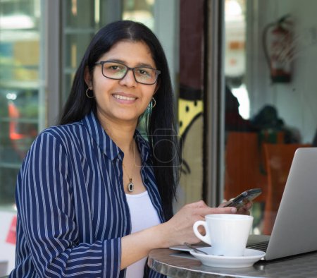Foto de Joven latina en café colombiano, trabajando con laptop y celular en una tarde soleada - Imagen libre de derechos