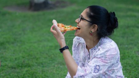 Foto de Mujer joven riendo y disfrutando de una rebanada de pizza en el parque. - Imagen libre de derechos