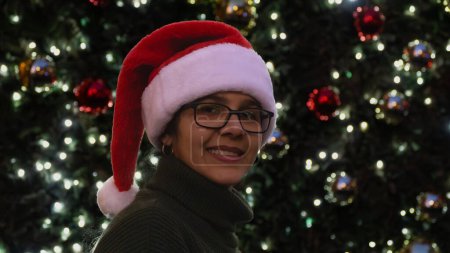 Foto de Mujer con lente sonriente y alegre, con sombrero de Santa y suéter verde de pie frente al árbol de Navidad con luces. - Imagen libre de derechos