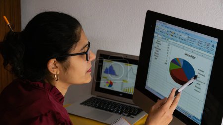 Foto de Mujer joven teletrabajo desde casa, sentada frente a una computadora analizando gráficos estadísticos. - Imagen libre de derechos