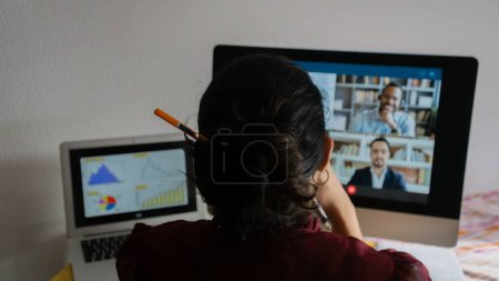 Foto de Mujer joven teletrabajando desde casa, sentada frente a una computadora con gafas y blusa de tinte de vino en una reunión de trabajo con agentes bancarios. - Imagen libre de derechos