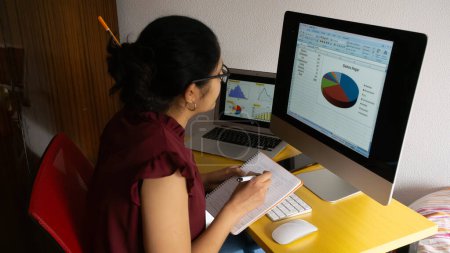 Foto de Joven latina frente a una computadora tomando notas de un análisis estadístico bancario con una blusa de vino tinto y copas. - Imagen libre de derechos