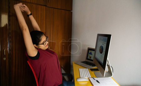 Foto de Mujer joven teletrabajo desde casa, sentada frente a un ordenador con gafas y una blusa roja estirándose. - Imagen libre de derechos