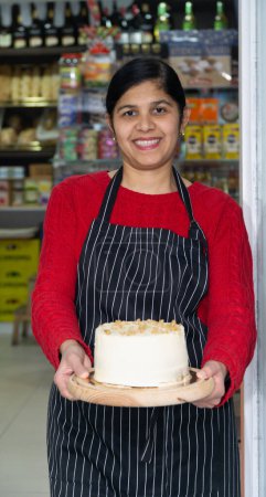 Foto de Joven emprendedora sonriente en delantal, sosteniendo pastel en la entrada del café - Imagen libre de derechos