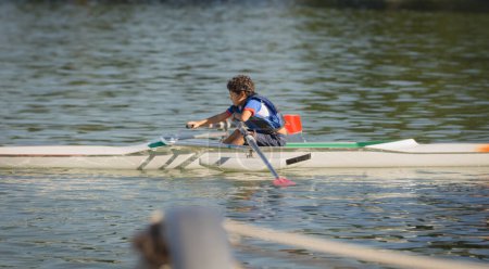 10 year old boy training rowing on a lake near his home with blue life jacket on a summer afternoon at a park lake.