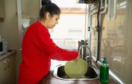 Foto de Una mujer en un suéter rojo está lavando un plato en un fregadero. El fregadero está junto a una botella de jabón para platos. - Imagen libre de derechos