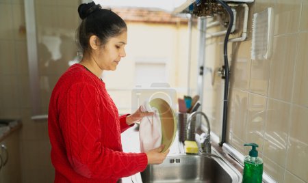 Foto de Una mujer está lavando un plato en un fregadero de cocina. Ella está usando un suéter rojo y se centra en su tarea - Imagen libre de derechos