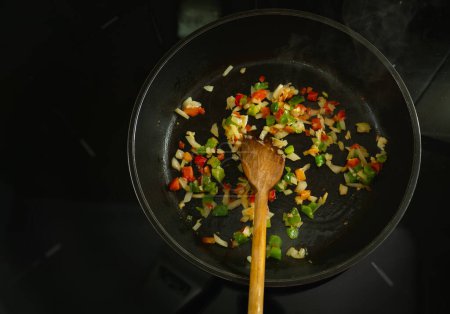 Foto de Se está cocinando una sartén de comida con una cuchara de madera. La comida es una mezcla de verduras, incluyendo pimientos y cebollas. La escena es cálida y acogedora - Imagen libre de derechos