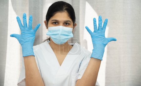 Foto de Una mujer con una bata blanca lleva guantes azules y una máscara. Ella sostiene sus manos en el aire - Imagen libre de derechos