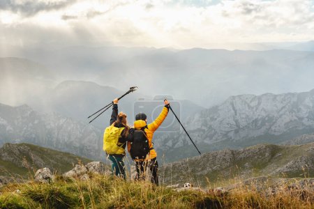 Téléchargez les photos : Quelques randonneurs avec des sacs à dos levant leurs bâtons de trekking alors qu'ils atteignent le sommet d'une montagne. alpinistes contemplant le paysage de la chaîne de montagnes. sports de plein air - en image libre de droit
