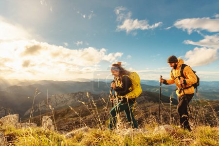 Téléchargez les photos : Deux alpinistes, hommes et femmes, montant une montagne au coucher du soleil. randonneurs équipés de sacs à dos, bâtons de trekking et vêtements chauds marchant dans la montagne. - en image libre de droit