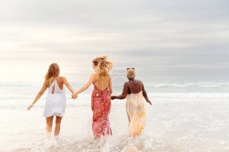 three young women holding hands walking towards the sea on a summer's day