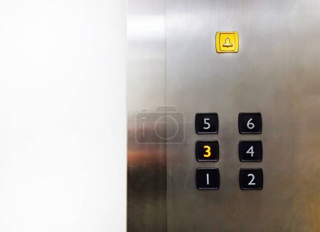 Grupo de pulsadores Black Elevator con campana amarilla, botones de advertencia o alarma sobre fondo de acero inoxidable con espacio de copia que muestra al pasajero presionado en el tercer piso para subir o bajar.