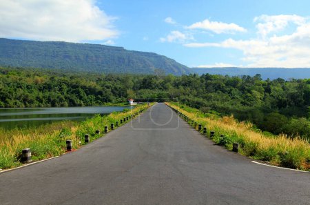 The road stretches along a beautiful river or lake, with mountains, blue sky, white cloud and green forests in the foreground at Wang Bon reservoir, Nakhon Nayok, Thailand.