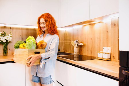 Téléchargez les photos : Joyeux jeune femme en bonne santé avec des légumes frais dans une boîte en plastique verte dans ses bras en souriant à la caméra dans leur cuisine. Photo de haute qualité - en image libre de droit