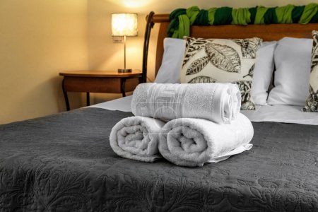 Primer plano de toallas blancas cuidadosamente enrolladas colocadas en una cama. Mesita de noche y almohadas proporcionan un telón de fondo borroso. Ideal para conceptos de hospitalidad, hotel e interiorismo. Enfoque selectivo.