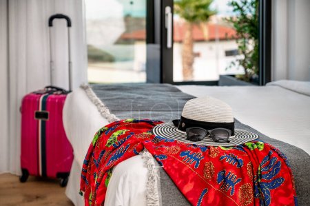 Téléchargez les photos : Doté d'un chapeau débordant d'été et de lunettes de soleil disposées sur le bord d'un lit, complétées par un paréo coloré. En arrière-plan flou, une valise ajoute à l'anticipation d'un voyage. - en image libre de droit