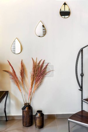 Cozy home decorative detail featuring three drop-like small mirrors adorning the wall, complemented by metal vases filled with dried florals, adding warmth and charm to the interior space.