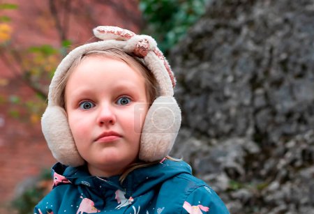 Téléchargez les photos : Portrait d'automne d'une fille de 7-8 ans, aux yeux grands ouverts surpris, dans des écouteurs chauds et moelleux - en image libre de droit