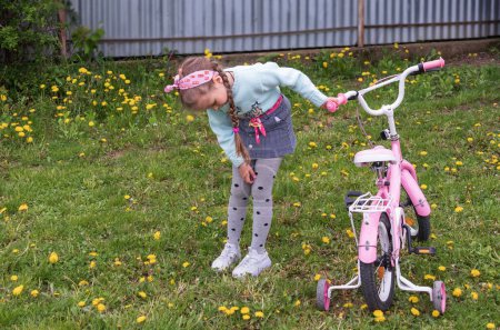 Photo for A little girl stands in the yard on green grass with dandelions and carefully examines a children's bicycle - Royalty Free Image