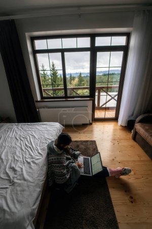 Téléchargez les photos : Jeune femme occupée avec ordinateur portable assis sur le sol par lit double et réseautage ou tout simplement passer du temps libre sur divers sites Web - en image libre de droit