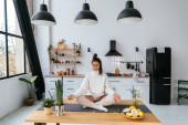 Shot of pretty young woman in lotus position sitting on the table while relaxing in the kitchen at home. Poster #625890796