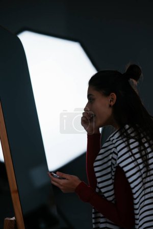 Téléchargez les photos : Joyeux jeune femme applique du maquillage sur son visage devant le miroir - en image libre de droit