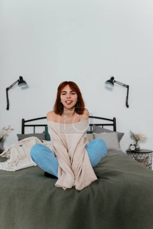 Foto de Mujer joven adulta con el pelo rojo en ropa casual sentada en la cama en casa - Imagen libre de derechos