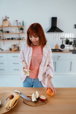 Foto de Feliz mujer sana cortando frutas en una tabla de madera mientras se hace el desayuno en una cocina - Imagen libre de derechos
