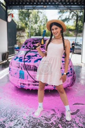 Téléchargez les photos : Young woman high pressure hose stands in front of a car covered in pink foam - en image libre de droit