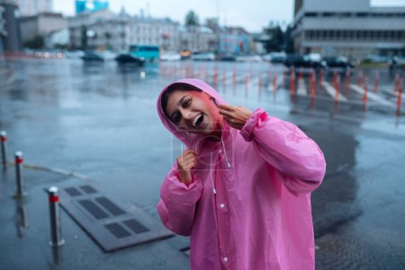 Téléchargez les photos : Young smiling woman with a pink raincoat on the street while enjoying a walk through the city on a rainy day. - en image libre de droit