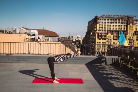 Foto de Young sporty happy woman in sportswear doing stretching exercises on yoga mat on house roof in early morning - Imagen libre de derechos