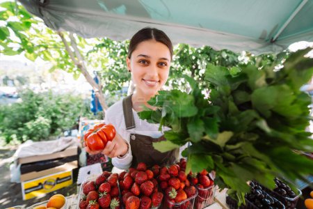 Téléchargez les photos : Young positive salesgirl at job, selling sells home-grown vegetables and fruits in vegetable shop - en image libre de droit