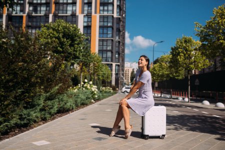 Téléchargez les photos : Jolie jeune femme assise sur une valise dehors. Attrayant voyageur sourit dans la rue. - en image libre de droit