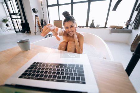 Téléchargez les photos : Jeune femme travaillant sur un ordinateur portable tout en prenant une baignoire à la maison - en image libre de droit