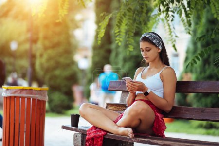 Foto de Chica feliz usando un teléfono inteligente en un parque de la ciudad sentado en un banco - Imagen libre de derechos