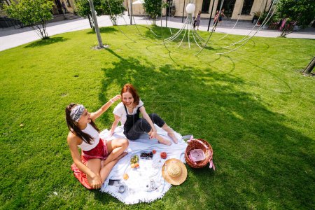 Foto de Two women having picnic together, sitting on the plaid on the lawn - Imagen libre de derechos