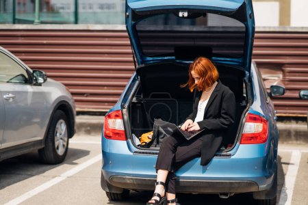 Téléchargez les photos : Affaires attrayant jeune femme travaillant sur ordinateur portable tout en étant assis dans le coffre de la voiture tout en voyageant - en image libre de droit